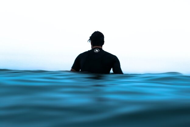 Ein Mann im Neoprenanzug steht im Meer, er ist von hinten zu sehen